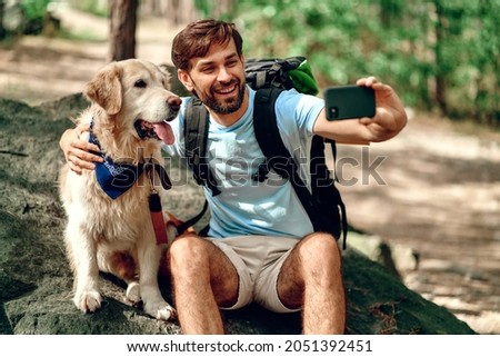 A male hiker with a backpack and a labrador dog makes a selfie on the phone while sitting on a stone in the forest. Camping, travel, hiking.