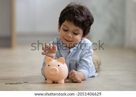 Happy small adorable mixed race child boy putting coins in piggybank, learning saving money for future purchase, calculating personal budget or managing finances, lying on warm heated floor. Royalty-Free Stock Photo #2051406629