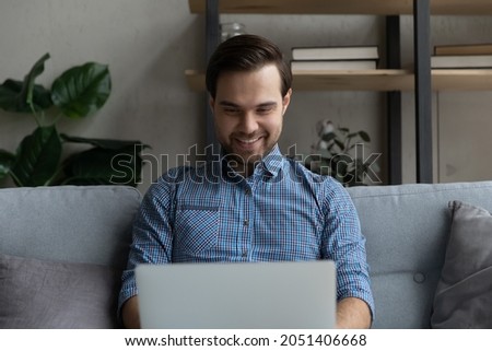 Smiling man using laptop, sitting on couch at home, happy young male looking at computer screen, spending leisure time with modern device, chatting with friends in social network, shopping online