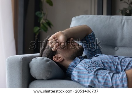 Sick man feeling headache migraine or dizziness, covering eyes with hand, lying on couch at home, exhausted tired millennial male sleeping after difficult working day, resting on comfortable sofa Royalty-Free Stock Photo #2051406737