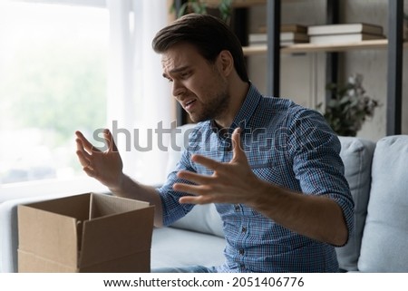 Unhappy man frustrated by wrong order online shopping or damaged package, bad delivery service, upset confused Caucasian customer opening unpacking parcel, cardboard box, sitting on couch at home Royalty-Free Stock Photo #2051406776
