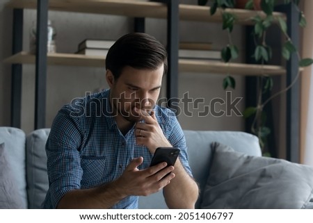 Thoughtful serious man touching chin, looking at smartphone screen, sitting on couch at home, pensive young businessman or student reading bad news in message, unexpected debt, solving problem Royalty-Free Stock Photo #2051406797