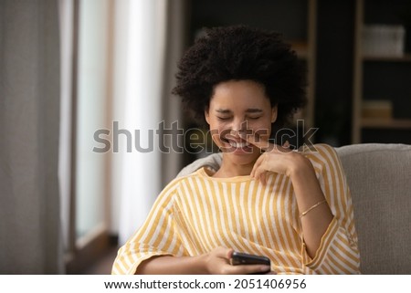 Laughing millennial generation mixed race african ethnicity woman holding smartphone in hands, watching entertaining video or photo content, reading message with good news, having fun at home.