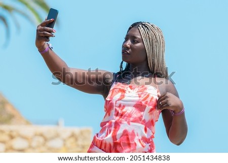 Calm african woman with braids and colorful dress taking a selfie on a sunny day with a clear sky background outdoors  Selective focus 