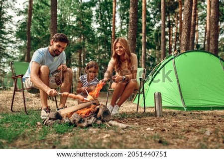 Happy family with a child on a picnic sit by the fire near the tent and grill a barbecue in a pine forest. Camping, recreation, hiking. Royalty-Free Stock Photo #2051440751