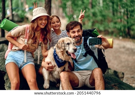 Mom and dad with their daughter with backpacks and a labrador dog take a selfie on the phone while sitting on a stone in the forest. Camping, travel, hiking.