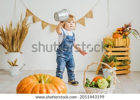 A little girl plays with a mini bucket in the autumn harvest. Little girl in denim overalls next to a large pumpkin. 