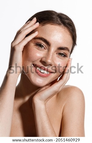Vertical portrait of beautiful girl smiling at camera, touching natural face without makeup, healthy glowing facial skin and white teeth, standing over white background