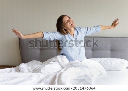Happy teen millennial girl feeling joy, full of energy after sleep enough, awaking. Cheerful young woman sitting in bed on comfortable mattress, stretching hands, body, smiling with closed eyes Royalty-Free Stock Photo #2051476064