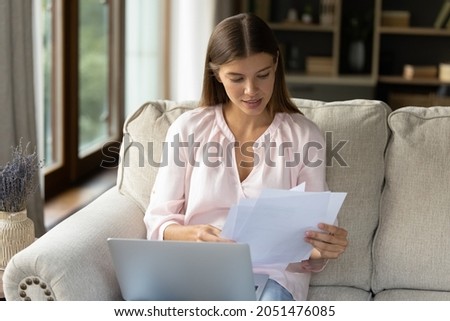 Focused young female homeowner studying paper bills, insurance, mortgage, loan agreement from bank, doing paperwork at home. Student receiving, reading document from school, college at laptop Royalty-Free Stock Photo #2051476085