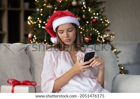 Sad thoughtful Xmas girl typing text message on smartphone, chatting online, resting on couch at gift box, Christmas tree. Woman with mobile phone feeling sad, frustrated, lonely at New Year night