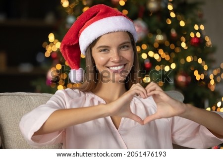 Happy girl in red Santa assistant hat making hand heart shape, showing love symbol, looking at camera, smiling. Young woman promoting Christmas Noel giveaway support, help. Head shot portrait