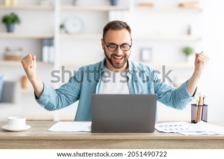 Young Caucasian guy gesturing YES in front of laptop, celebrating success at home office. Millennial man working with portable pc, making great deal, signing online contract, excited over achievement