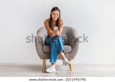 Happy Caucasian woman using cellphone, chatting on internet, working or studying online, sitting in armchair against white studio wall, full length. Cheery young lady watching video on mobile phone Royalty-Free Stock Photo #2051490740