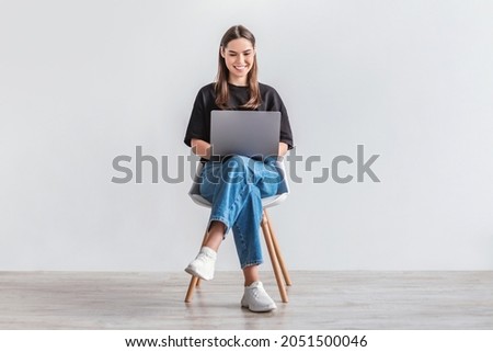 Happy young woman working online, sitting on chair and using laptop against white studio wall, full length. Positive Caucasian lady surfing internet, watching webinar on portable computer Royalty-Free Stock Photo #2051500046