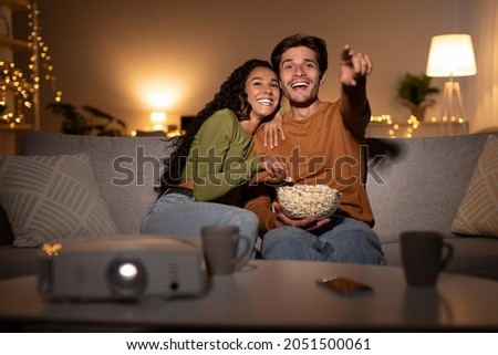 Joyful Couple Watching Film Online Using Home Cinema Projector Enjoying Evening Together On Weekend Sitting On Sofa In Living Room Indoors. Family Leisure, Entertainment And Fun Concept