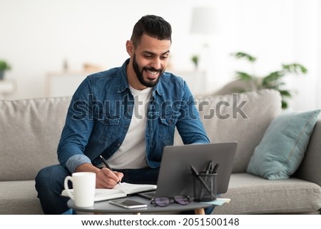 Happy young Arab guy working or studying on laptop computer, taking notes, having business meeting indoors. Millennial Eastern freelancer having video conference or webinar at home