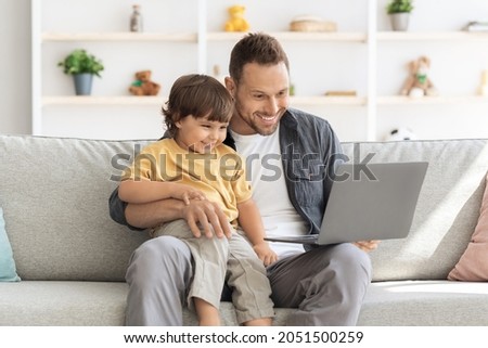 Early education and kids development. Loving father watching together cartoon on laptop to his cute little son, sitting together on sofa at home, enjoying time together