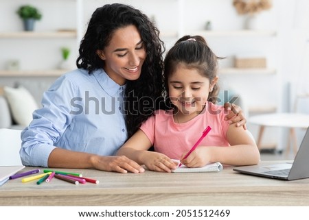 Caring arab mother helping her little daughter with homework and study while sitting together at desk in living room, cute small girl writing or drawing in front of laptop at home, copy space
