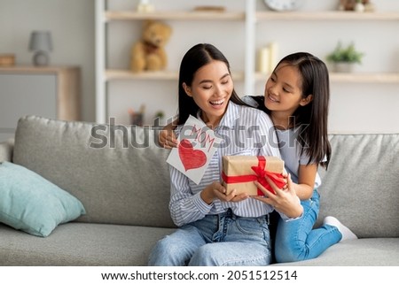 Congratulations. Loving asian daughter greeting surprised mom with Mother's Day, giving her handmade card and gift box, sitting on sofa at home interior, free space Royalty-Free Stock Photo #2051512514
