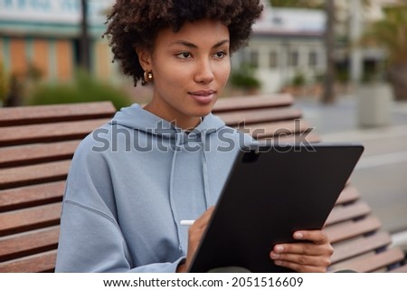 Creative serious curly haired woman draws sketches in tablet with stylus makes picture works on future project dressed in sweatshirt poses on wooden bench against city background uses modern device