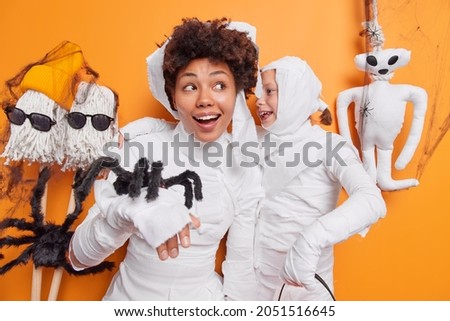 Small glad child whispers secret to mommy dressed like scarying mummies have fun together at halloween party surrounded by spooky characters isolated over orange studio background. Horror theme.
