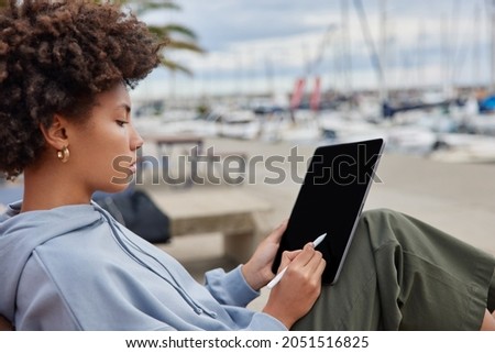 Sideways shot of professional female retoucher edits photos with stylus holds modern tablet dressed casually poses on pier admires beautiful sea view with yachts and boats. Woman graphic designer