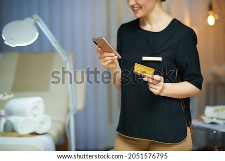 Closeup on smiling woman worker with smartphone and credit card browsing online shop in modern beauty salon.