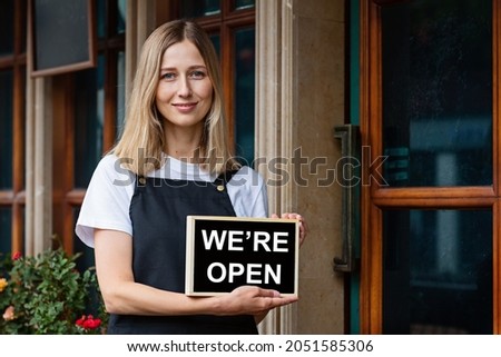 Young caucasian woman 30 years old with short blonde hair holding open signboard near doors of cafe. Reopen restaurant after coronavirus covid-19 quarantine shutdown. Welcome to cafeteria 