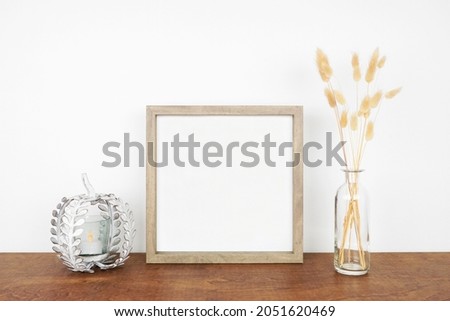 Mock up square wooden frame with fall grasses and pumpkin candle holder on a wood shelf against a white wall. Autumn concept. Copy space.