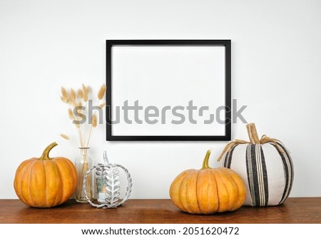 Mock up black frame hanging from a white wall with pumpkins and fall decor on a wood shelf. Autumn concept. Copy space.