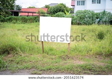 Blank white empty mockup template of a real estate sign at front of a lot of vacant land in a suburban neighbourhood with some residential houses at the background.