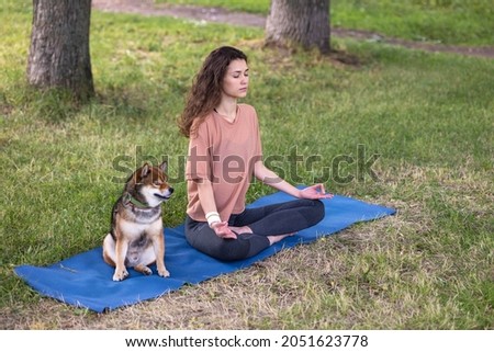 a sporty European woman does yoga and meditation in a park or a public place. a woman does sports with her pet outdoors. mental and physical health, physical exercises and breathing practices for Royalty-Free Stock Photo #2051623778