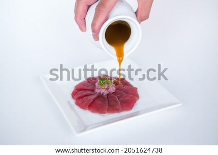 Hand pouring sauce on Tuna sashimi on white plate on white background. Isolated on white. The process of making and decorating tuna sashimi. Cooking
