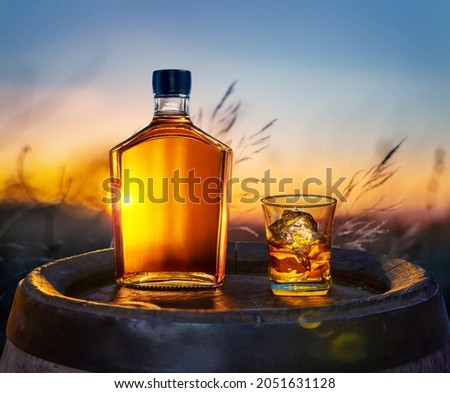 Carafe of whisky and glass of whisky with ice cubes on old wooden cask. Beautiful sunset at the background. Royalty-Free Stock Photo #2051631128