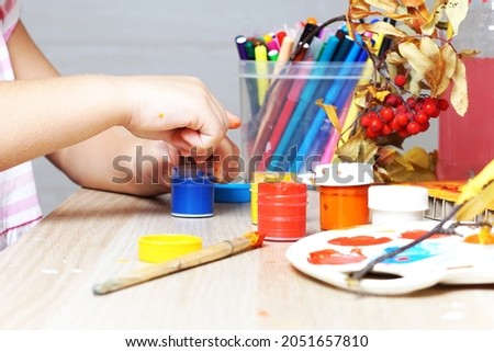 The child's hands of the girl dip the object in blue paint. In the background, red rowan and felt-tip pens. Paints on the table. The child draws and makes crafts in the kindergarten.