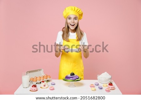 Teen fun girl chef cook confectioner baker in yellow apron cap at table taking photo of dessert by mobile cell phone isolated on pastel pink background studio Mousse cake food process workshop concept