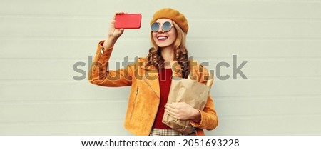 Portrait of happy smiling woman taking a selfie by smartphone holding grocery shopping paper bag with long white bread wearing an yellow beret on gray background
