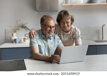 Happy middle aged senior family couple using computer apps, having fun web surfing useful information, shopping in internet store, communicating distantly, spending time online in modern kitchen.