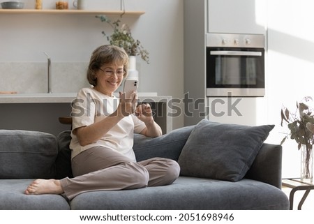 Happy old mature woman in eyewear using smartphone applications, resting on cozy sofa at home. Smiling middle aged female pensioner holding video call conversation on cellphone or shopping online. Royalty-Free Stock Photo #2051698946