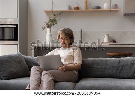 Happy relaxed middle aged 50s woman in eyeglasses using computer software applications, sitting on cozy couch. Smiling old mature grandmother enjoying communicating online, shopping or web surfing.