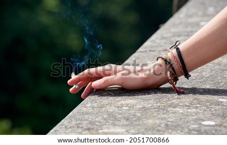 A female's hand wearing bracelets leaning on a stone structure holding a lit cigarette