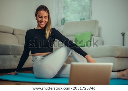 Attractive young woman using laptop while training at home and sitting on a fitness mat