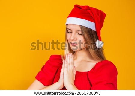 Young beautiful woman wearing Santa Claus Christmas hat over isolated yellow background, praying with hands together asking for forgiveness, smiling confidently.