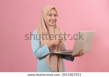 Portrait of a smiling young beautiful muslim girl holding laptop computer and pointing isolated over pink background.
