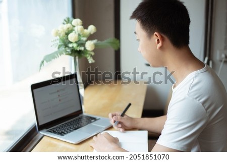 Online studying concept the young man in simple white T-shirt and black trousers working by using his laptop to search information according to the brief concepts written down on the notebook.