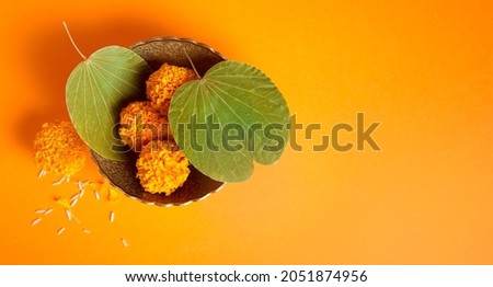 Dussehra greeting card photo background with clear space for message. Top view of a Dussehra leaves and marigold flower in a bowl on saffron color background. Royalty-Free Stock Photo #2051874956