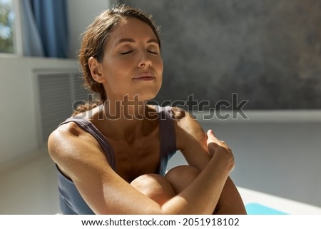 Close-up indoor image of good-looking Caucasian female taking deep breath with enjoyment, sitting with eyes closed, arms holding bent knees, relaxing and meditating at sport studio Royalty-Free Stock Photo #2051918102