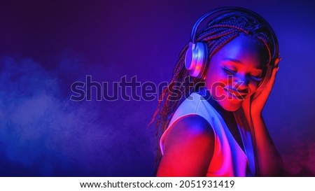 Stylish fashion african american teenager model wearing headphones listening dj music dancing in purple neon lights. Young teen girl enjoy cool music Royalty-Free Stock Photo #2051931419