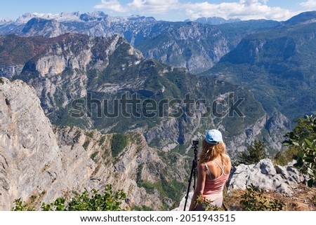 A young woman traveler stands on top of a mountain looking down at the Grlo Sokolovo Gorge and the surrounding mountains, next to an action camera on a tripod, capturing a breathtaking time-lapse view